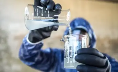 Imagem ilustrativa de banco de imagens: técnico de laboratório com traje de proteção despejando água entre dois béqueres