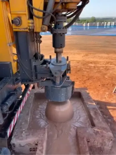 Close da cabeça de perfuração descendo na boca do poço, envolta em lama de perfuração dentro da caixa de concreto, com terra vermelha ao fundo