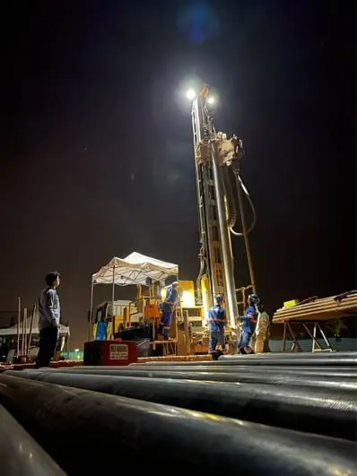 Perfuração à noite: a torre da sonda iluminada por um refletor no alto do mastro, quatro técnicos de uniforme ao pé dela e tubos de revestimento empilhados em primeiro plano