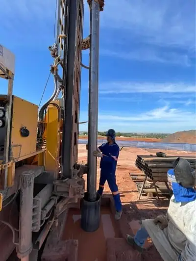 Técnico de uniforme azul e capacete operando a sonda ao lado da haste de perfuração, sob céu azul